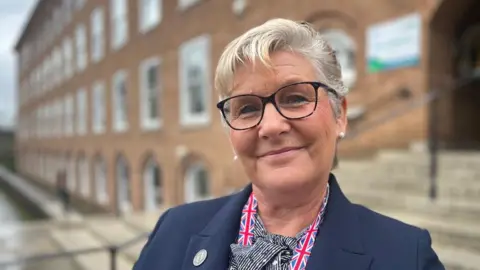 Jacqueline Fry wearing tortoiseshell-framed glasses, a black and white top and a navy blue blazer with a Union Flag lanyard and standing in front of County Hall in Exeter