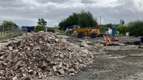 a pile of rubble in the foreground, with a dump truck, tractor and lorry in the background