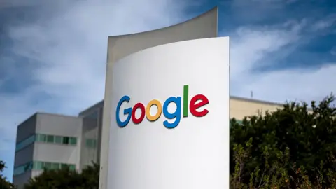 Google's multi-coloured logo is seen on a white sign in outside their headquarters, a grey and beige building with trees in the foreground and wispy clouds against a blue sky, in Mountain View, California in 2024