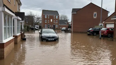A car is stranded in floodwater in a road through a housing estate. The water is a muddy brown and reaches up to the lower walls of the houses