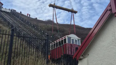 Redcar & Cleveland Borough Council A red and white carriage sits at the bottom of the tram tracks, which runs up the steep bank of the Saltburn cliffs. Harnesses have been attached to its underside and it is about to be lifted from the tracks by a crane. A crowd of people look down from the top of the cliffs.