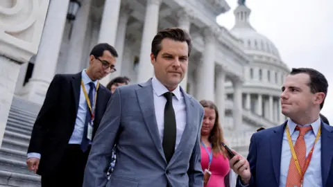 Reuters Matt Gaetz dalam setelan abu-abu dengan dasi gelap, di depan gedung marmer putih, dengan wartawan di sekelilingnya