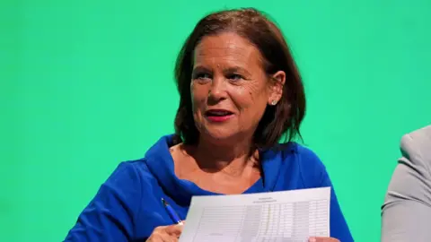 PA Media Sinn Féin leader Mary Lou McDonald at the Sinn Féin Ard Fheis. She has shoulder length brown hair and is wearing a bright blue top. The background behind her is green.