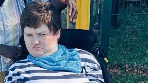 Lesley Honey-Green Jeni Honey-Green with short, dark brown hair sits in a wheelchair outdoors near a fenced area, with another person standing behind. A sign reading “Veolia Environmental Trust” is visible in the background, along with grass, a paved path and nearby buildings.
