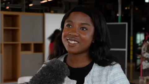 Young person being interviewed, smiling to camera in front of a microphone, with black hair.