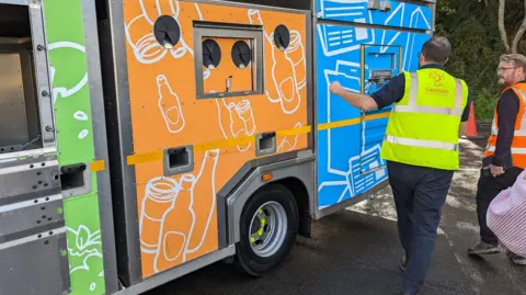 Two men wearing hi-vis waistcoats next to a recycling truck. One is holding a bag, while the other is putting something in the van.