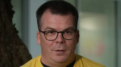 A man with black glasses and a yellow jumper looks at the camera. He has black hair