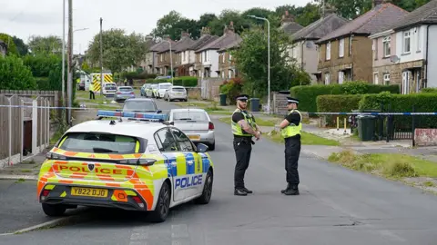 PA Media Two police officers stand next to a police car alongside police tape cordoning off Westbury Road in Bradford, with houses bordering the street in the background, on 21 August