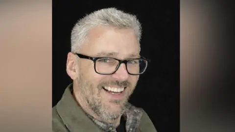 Craig Hurcombe smiles at the camera. He has short grey hair and is wearing black, thick-rimmed glasses and a brown shirt. He is pictured against a black background.