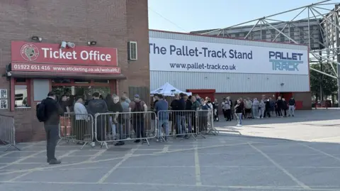 The word ticket office is above windows on the outside of the stadium on the left of the photo. The queue starts there, with people behind barriers, and continues towards the right of the image outside by the club's main stand.