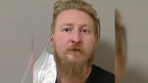 Oakes has ginger short hair, a moustached and a log beard. he has blue eyes. He is staring at the camera as a mask hangs from his ear.