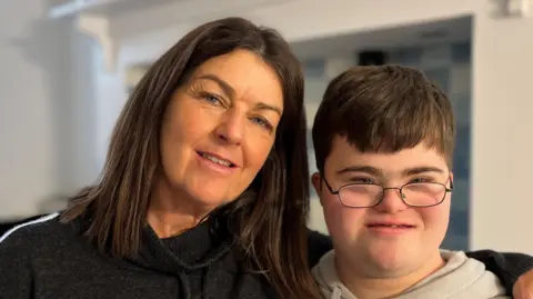 A woman with shoulder length brown hair, wearing a black top, has her arm around a boy who has short brown hair and glasses