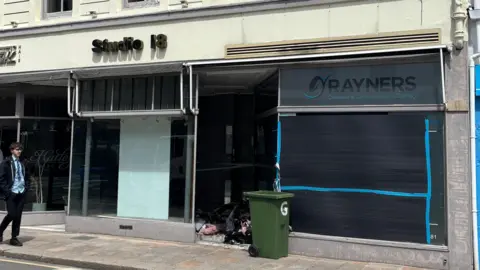 The front of a dry cleaners which has been boarded up after a fire. The dry cleaners is called Rayners and is next to a hair salon. A green wheelie bin is outside the shop. A boy wearing a school uniform is walking along the street.