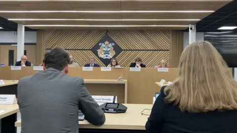 LDRS Councillors at the cabinet meeting of Stockton Council. The image is taken from the public gallery, with the back of two council officers - a man and a woman - in the foreground. The cabinet is sat opposite them at a long raised table, with their names displayed on a sing in front of each. Behind them is the emblem of the council, displayed on a herringbone patterned wooden panel. 
