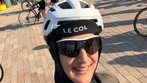 Shabana Seedat A woman smiles at the camera, wearing a helmet with a Le Col hat underneath. She is wearing cycling sunglasses and is standing on a brick pavement surrounded by bikes and cyclists