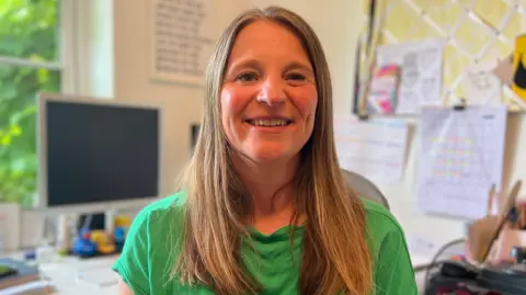 A woman with long hair, wearing a green shirt, is seated in an office setting. Behind them is a desk cluttered with a computer monitor, papers, and office supplies. A window on the left reveals greenery outside. The wall in the background features pinned papers and a framed text print.