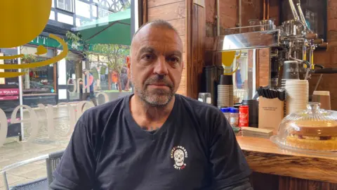 BBC Pete Masters, wearing a blue tshirt, sat in front of the wooden counter of his cafe in Hereford, with a window showing the street behind him