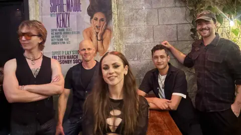 Sophie-Maude Sophie-Maud is pictured standing in front of her band. She is wearing a black top with holes in the front and she has long brown hair. There are four other band members - all of them male and all of them wearing dark-coloured clothing. Two are sitting at a wooden table and the other two are standing. Behind them, on a wall, is a poster of the band.