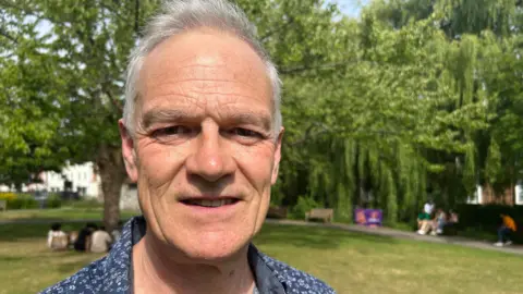 White man with white grey hair wearing a blue floral shirt looks at the camera. Behind him is a park with green lawn and a weeping willow tree'