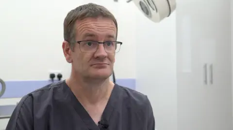 PCN director Dr Rob Caudwell in dark grey surgical scrubs, wearing glasses with slightly greying hair