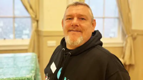 A man with grey hair and a grey beard, wearing a dark hoodie, looks at the camera
