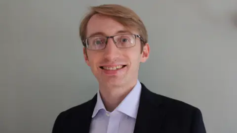 RoSPA A head and shoulders photo of James Broun from RoSPA smiling at the camera. He has short, fair hair and glasses. He is wearing a black jacket and an open neck lilac shirt.