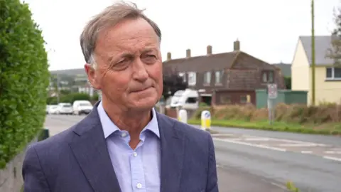 A picture of Andrew wearing a purple suit shirt and a navy colour blazer. He has brown and grey hair and is stood in front of a road.