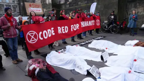 PA Media Protesters at the Court of Session in Edinburgh. Some are holding a big banner saying CODE RED FOR HUMANITY while several others are lying on the ground with white sheets over them, as if they are corpses (presumably killed by the climate crisis).