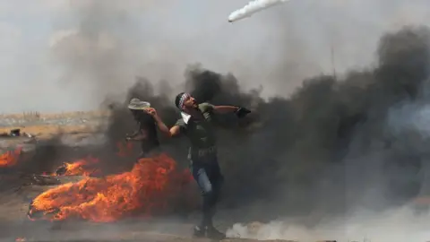 Reuters A Palestinian man uses a tennis racquet to return a tear-has canister fired by Israeli troops during a protest near the Gaza-Israel border fence (11 May 2018)