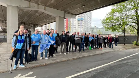 Marian McNamee/BBC About 30 Coventry City fans, some holding flags, are standing in White Street coach park waiting to be transported to Blackburn