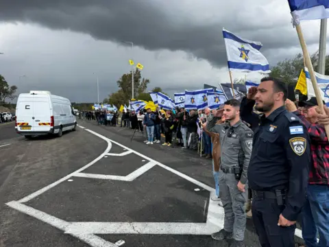 Israel Police Police officers line the street holding flags as a white van drives by under a grey, cloudy sky