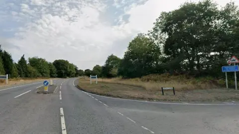 A Google street view image of a road junction. A single carriageway can bee seen with traffic island in the centre and two white signposts either side on grassy verges, lined by trees. A turning to the right is signed Rocky Lane and a sign warns of people riding horses in the area.