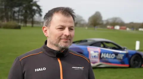 Qays Najm/BBC Paul Hamilton is standing in a grassy area, wearing a black and orange zip-up top. In the background, slightly blurred, is a blue McLaren car, emblazoned with the Ipswich Town badge and the Halo logo. 