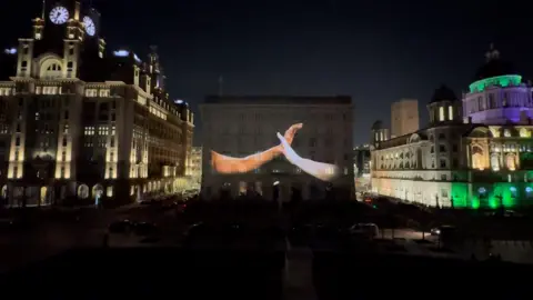 Two hands come together, illuminated on a large building, at night time, on a waterfront