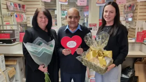 An elderly man stands between two women who hold flowers and gifts. He holds a red cardboard heart which reads 'post office'. 