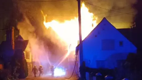 Firefighters spray water from hoses on to a house, which has flames pouring out of it and smoke.