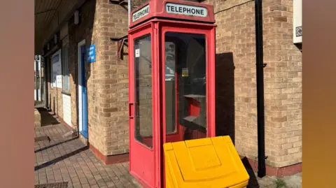 A traditional red telephone box positioned on a brick-paved walkway beside a building made of tan-colored bricks. The telephone box has glass panels on all sides, and the word “TELEPHONE” appears in white lettering on a sign at the top of the box on at least two visible sides. The paint on the box appears worn in some areas.