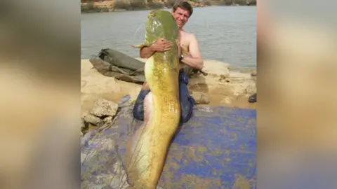 Simon Bartlam/WBC Simon Bartlam, kneeling topless on the side of water, holding a huge green fish which he has caught.
