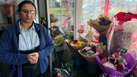 A woman wearing a blue cardigan and blue shirt with white stripes stood next to bouquets of flowers. She is in the front of her shop and the glass window shows the street in front of her shop.