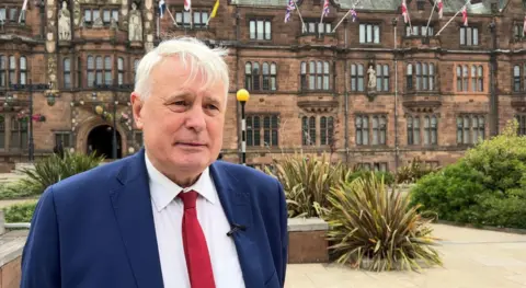 A man stands outside an ornately decorated council house. He wears a dark blue suit, white shirt and red tie.