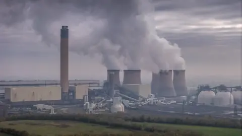 Getty Image Drakes Power Station. The photo features smoke billing from six stout chimney. The sky is cloud and smoke colored. There are trees and grasses in the foreground.