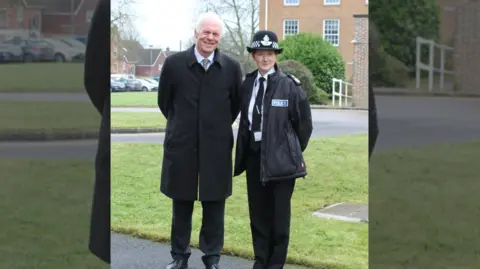 Wiltshire Police two people standing next to each other looking at the camera in front of a grassy verge with buildings in the background. The woman on the right wears a police hat, coat and tie, while the man on the left wears a heavy overcoat