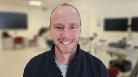 Mark is wearing a black jacket and is smiling at the camera. He has a shaved head and some facial stubble. The background is blurred but you can see there a big computer screens in the lab behind him.