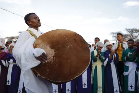 Anadolu via Getty Images A traditional drummer bangs his instrument as people clap along in the background.