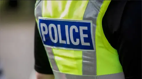 A general image of the back of an unidentified police officer. They are wearing a black shirt. On top they are wearing a high-vis green vest with a blue badge in the centre which reads 'police' in white writing.