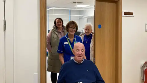 Jim Ross wearing a white and blue checked shirt under a blue jumper being pushed through a door in a wheelchair by nurse Michelle Doherty. His wife Lorna wearing a blue jumper, a beige waterproof hooded coat and a gold necklace and his daughter Gayle wearing a pink hoodie with a beige hood bodywarmer can be seen in the room behind the nurse through an open door. 