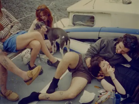 Kuba Ryniewicz A group of young adults on a small boat. On the right a man and woman are lying down, the woman with her head on the man's stomach. They both have brown short hair and are looking at each other in mid-conversation. There are collection of lighters next to them as well as an empty bottle. To the left there is a woman with red hair stroking a grey and white whippet. The legs of two other people are visible on the left, both wearing shorts.