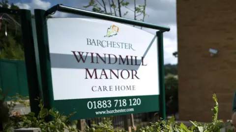 A white sign outside a building reading BARCHESTER WINDMILL MANOR CARE HOME 01883 718 120