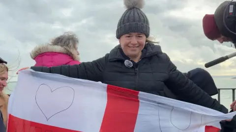 Helen wears a bobblehat and winter coat. She holds a St George's flag.