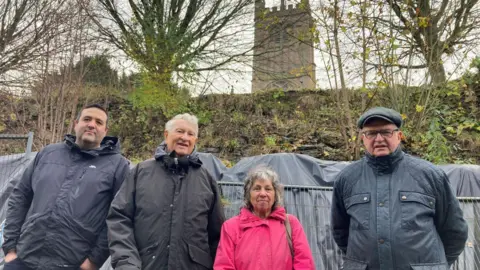 BBC Four people standing in front of metal barriers and granite-filled bags, supporting a collapsed stone wall. St Laurence's Church tower is in the background. 